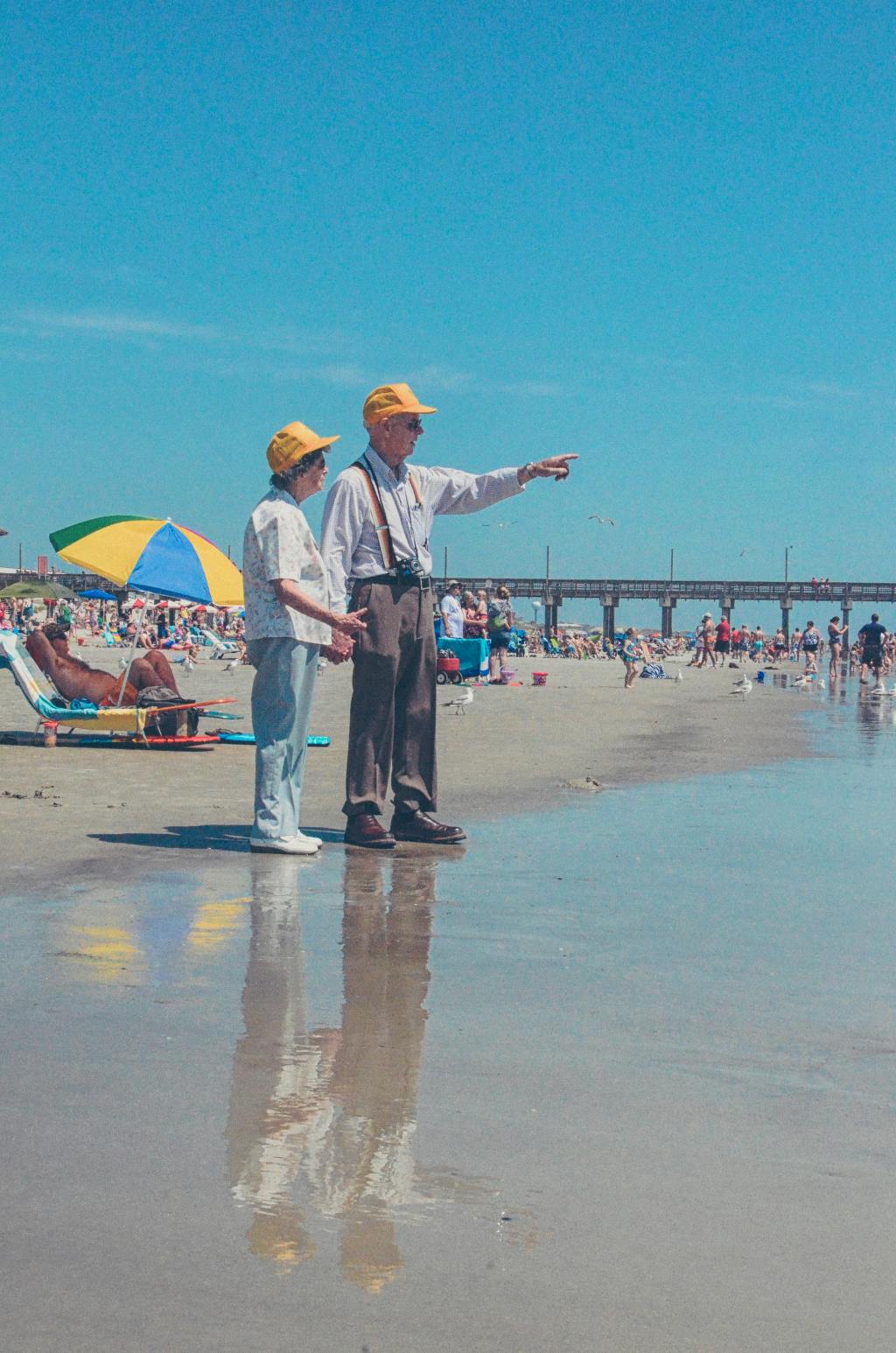 Deux personnes âgées sont à la plage, habillés et portant des chapeaux jaunes. Ils se donnent la main. L'homme pointe quelque chose vers l'horizon.